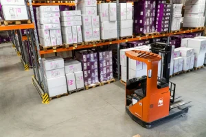 Un almacén con cajas apiladas en estantes metálicos y una carretilla elevadora naranja en primer plano, mostrando tecnología moderna de gestión de inventario, capturado en foto-1616401784845-180882ba9ba8.